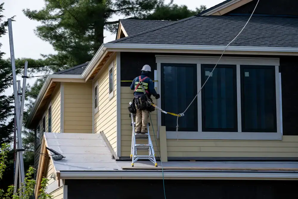 A construction worker in safety gear stands on a ladder placed on a roof, working on the exterior of a yellow house with large windows and dark trim—showcasing expert Home Remodeling Essex County, MA style. Trees are visible in the background.