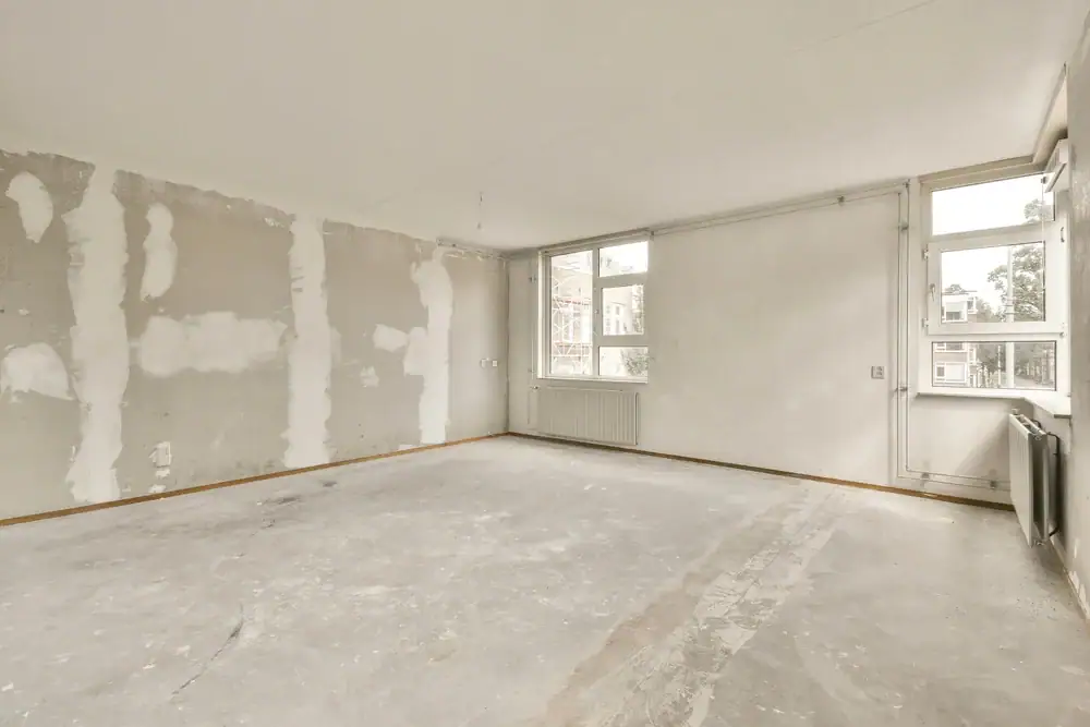 An empty, unfinished room with concrete floors and unpainted, patchy walls. Large windows on two walls let in natural light, with radiators below. Perfect for a Home Remodeling Essex County, MA project, the space appears to be under renovation.