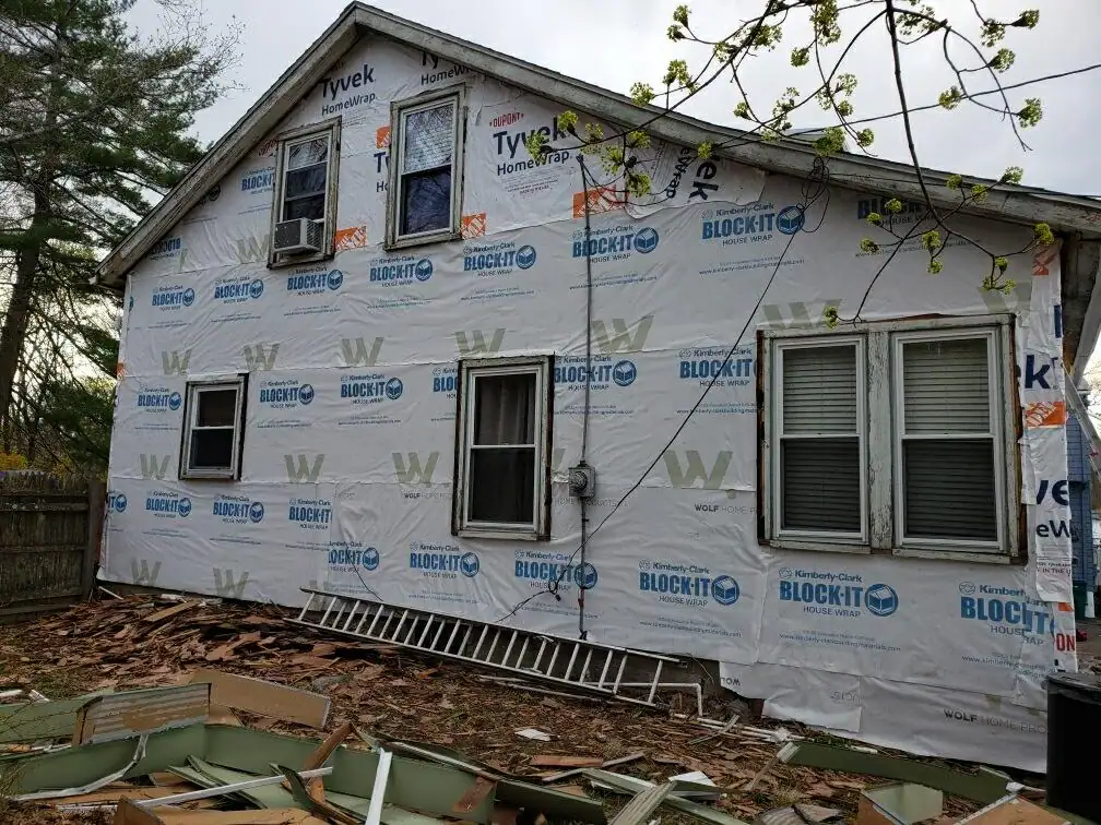 A house undergoing Home Remodeling in Essex County, MA, with exterior walls wrapped in white building material, surrounded by construction debris and a long ladder on the ground. Intact windows and trees are visible in the background.