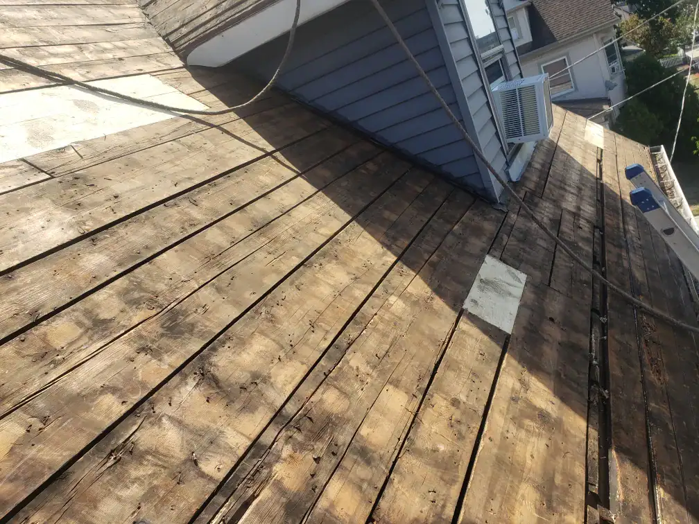Weathered wooden roof decking without shingles. A building rises above, featuring blue siding and a window air conditioner. Tools and other houses are visible in the background, showcasing typical Home Remodeling Essex County, MA projects.