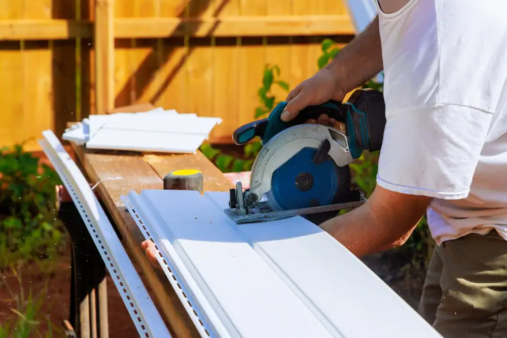 A person uses a circular saw to cut white vinyl siding on a workbench outdoors in MA, with construction materials and a tape measure nearby&mdash;a typical scene in Home Remodeling Essex County. A wooden fence and greenery are visible in the background.