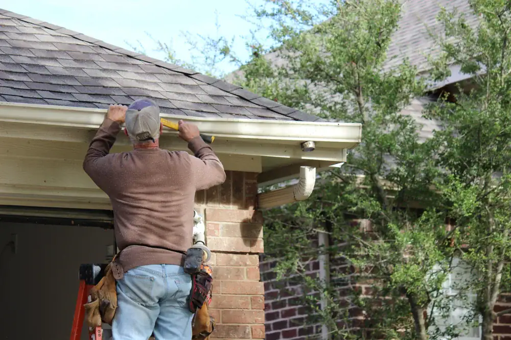 A man stands on a ladder fixing the gutter of a house, holding a level tool&mdash;a typical scene during home remodeling in Essex County, MA. He wears a long-sleeve shirt, jeans, cap, and tool belt, with trees and another roof visible in the background.